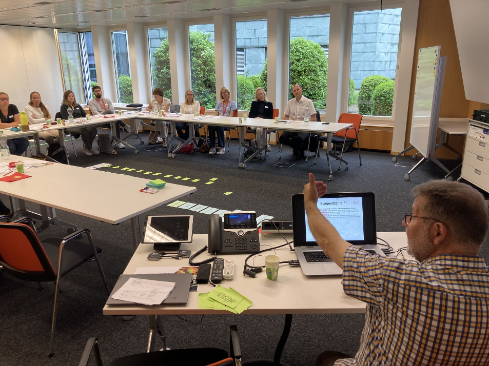 Seminarleiter zeigt eine Präsentation auf seinem Laptop, während er mit den Teilnehmenden in einem U-förmigen Tisch-Arrangement spricht. Auf dem Boden liegen beschriftete Moderationskarten von einer Gruppenarbeit.